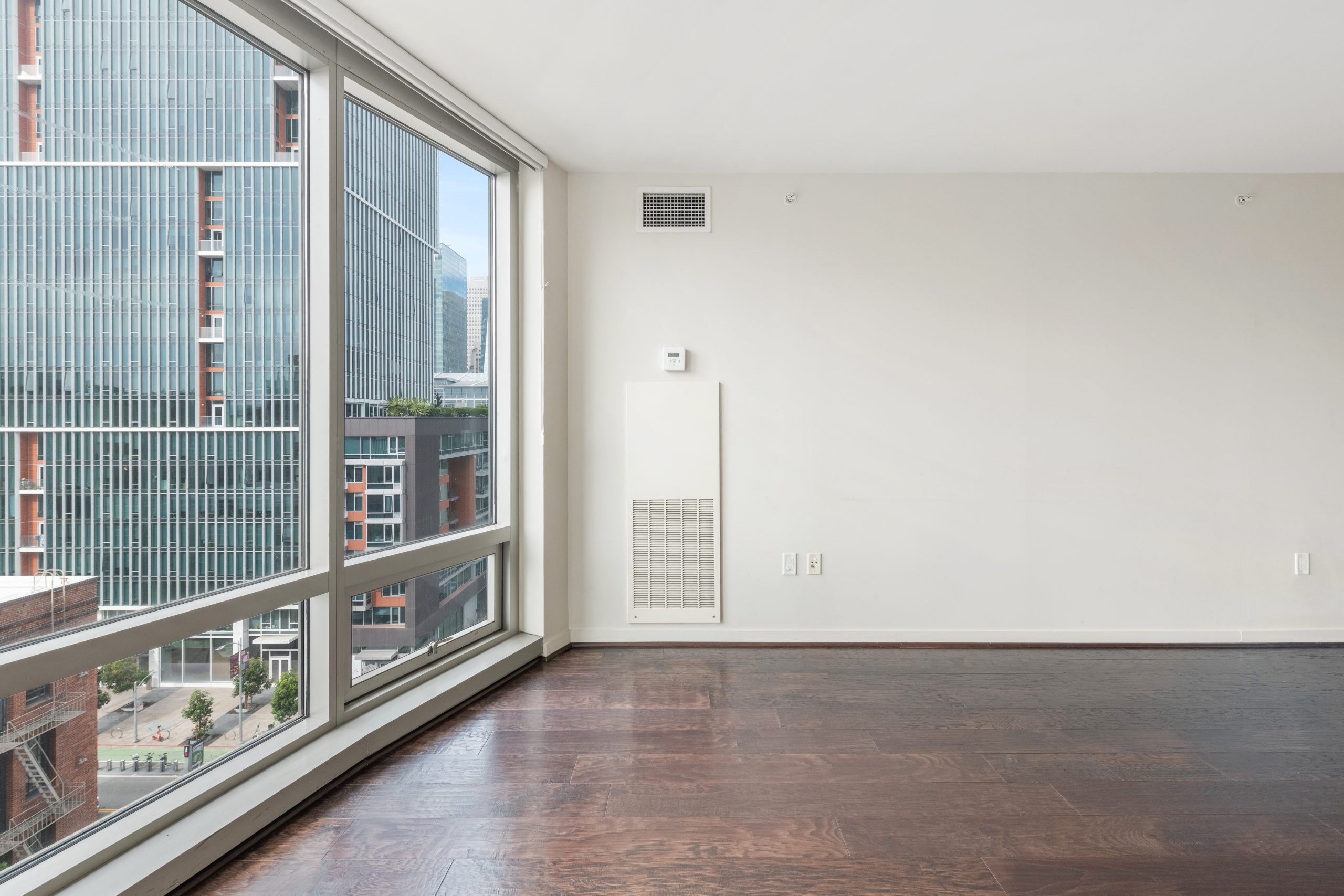 Open living room with dark hardwood floors and large windows overlooking neighboring buildings.